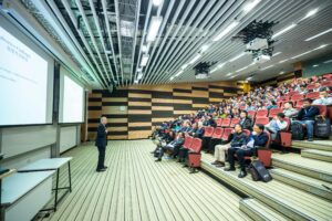 A speaker delivers a presentation to an attentive audience in a modern auditorium setting.